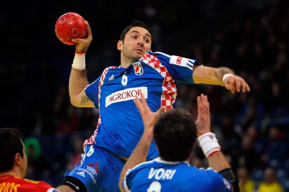 Ein Handballspieler in einem blau-rot karierten Trikot springt, um den Ball zu werfen, während zwei gegnerische Spieler in Blau und Gelb versuchen, ihn während eines Spiels zu blockieren.