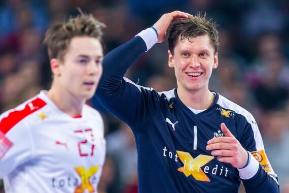 Zwei männliche Handballspieler auf einem Spielfeld, einer in einem dunkelblauen Trikot, lächelnd und den Kopf berührend, der andere in einem weißen Trikot mit der roten Nummer 27, beide mit sichtbaren Sponsorenlogos, Hintergrund unscharf.