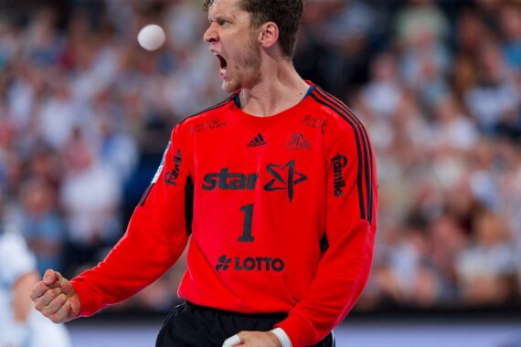 Ein männlicher Handballspieler im roten Trikot mit der Nummer 1 ballt während eines Spiels die Faust und jubelt, während im Hintergrund eine verschwommene Menschenmenge zu sehen ist.