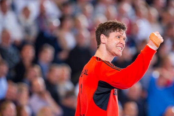 Ein männlicher Sportler in einem orange-schwarzen Trikot ballt die Faust und lächelt triumphierend, während er vor einer verschwommenen Menge in einer Sportarena steht.