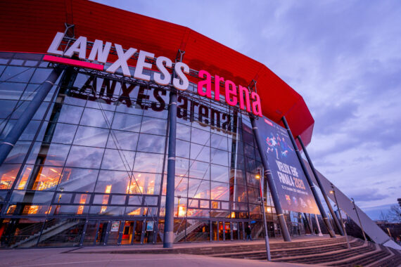 Das Äußere der LANXESS arena mit seinem großen roten Schild und der Glasfassade, in der sich die Lichter und der Himmel in der Abenddämmerung spiegeln. Veranstaltungsbanner und Logo sind auf dem Gebäude zu sehen.