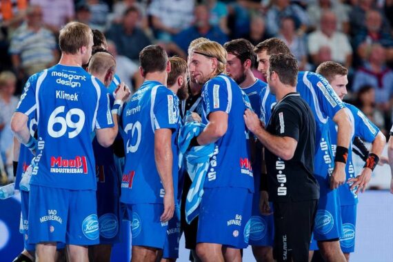 Eine Gruppe von männlichen Sportlern in blauen Trikots drängt sich auf einem Hallensportplatz zusammen, wahrscheinlich während einer Auszeit. Ein Trainer oder Mitarbeiter steht bei ihnen, und im Hintergrund ist eine Menschenmenge zu sehen.