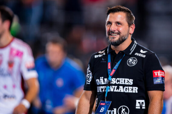 Ein lächelnder Mann in einem schwarzen Sporthemd mit Sponsorenlogos steht in einer Halle und trägt ein Schlüsselband und ein Abzeichen. Der Hintergrund ist unscharf und zeigt andere Menschen und eine Sportumgebung.