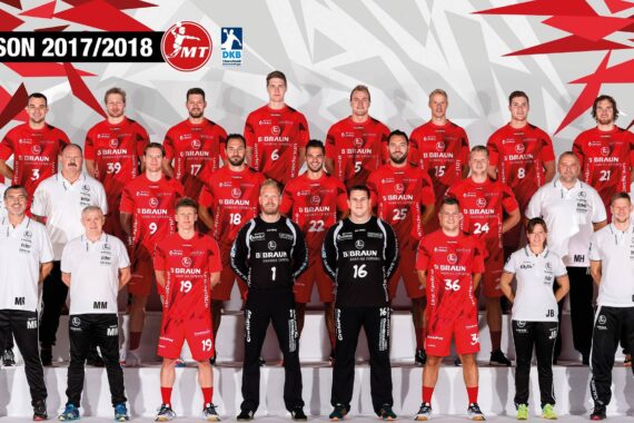 Eine Profi-Handballmannschaft in roten Trikots posiert für ein Gruppenfoto mit Trainern und Mitarbeitern in weißen Outfits. Der Hintergrund zeigt ein geometrisches rotes und weißes Muster mit SAISON 2017/2018 oben links.