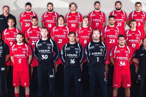 Eine Herren-Handballmannschaft stellt sich für ein Gruppenfoto auf. Die Spieler in roten Trikots stehen und knien in Reihen, während die Trainer und Betreuer in schwarzer Kleidung auf beiden Seiten und im Hintergrund stehen. Der Hintergrund ist ein neutrales, abstraktes Muster.