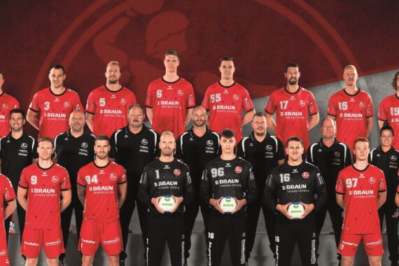 Eine Herren-Handballmannschaft in roten und schwarzen Trikots posiert für ein Gruppenfoto mit den in der Mitte stehenden Trainern. Die Spieler halten Handbälle in der Hand, und auf dem rot-grauen Hintergrund ist ein großes, schwaches Mannschaftslogo zu sehen.