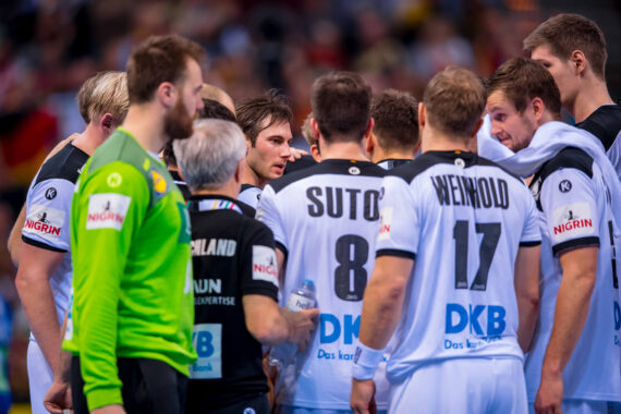 Eine Herrenhandballmannschaft sitzt während eines Spiels zusammen und diskutiert. Die Spieler tragen weiße Trikots mit schwarzer Umrandung, auf dem Rücken stehen Namen und Nummern. Die Atmosphäre ist intensiv und konzentriert.