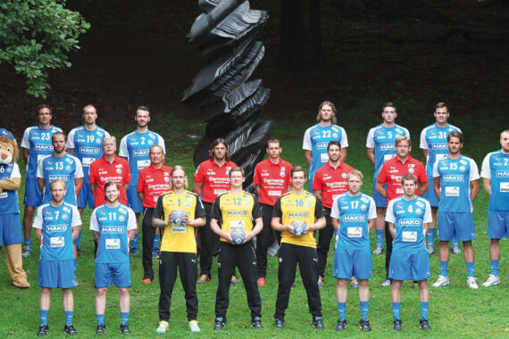 Eine Herrenhandballmannschaft posiert im Freien vor einer schwarzen abstrakten Skulptur. Die Spieler in blauen Trikots stehen neben einem Stab in Rot und zwei in Gelb, links steht ein Maskottchen im Löwenkostüm. Der Hintergrund ist von Bäumen geprägt.