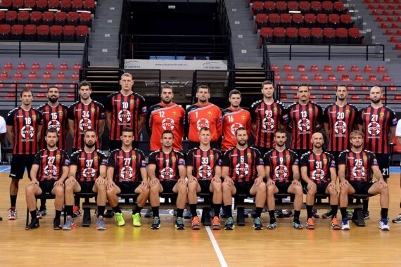 Eine Herren-Handballmannschaft posiert in einer Halle auf einem Spielfeld. Die Spieler in schwarz-rot gestreiften Trikots sitzen und stehen in zwei Reihen, flankiert von zwei Trainern in weißen Hemden. Im Hintergrund sind leere rote Stadionsitze zu sehen.