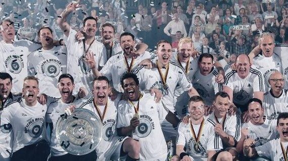 Eine jubelnde Fußballmannschaft in weißen Trikots feiert auf dem Spielfeld mit Medaillen und Trophäen, umgeben von Konfetti, während im Hintergrund eine jubelnde Menge zusieht.