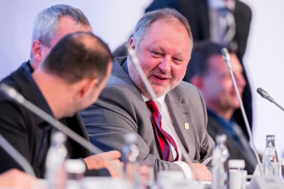 Eine Gruppe von Männern in Anzügen sitzt an einem Konferenztisch mit Mikrofonen und führt ein Gespräch. Der Mann in der Mitte, der einen grauen Anzug und eine rote Krawatte trägt, lächelt und schaut einen anderen Mann an. Auf dem Tisch stehen Wasserflaschen.