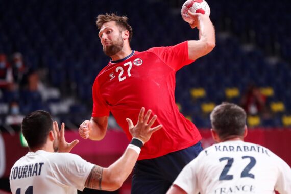 Ein Handballspieler in einem roten Trikot springt, um den Ball zu werfen, während zwei gegnerische Spieler in weißen Trikots versuchen, ihn während eines Spiels in einer Halle zu blockieren.