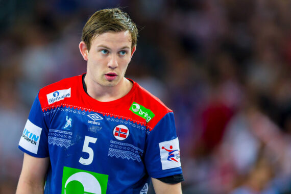 Ein männlicher Handballspieler in einem blauen Trikot mit der Nummer 5 und der norwegischen Flagge steht auf dem Spielfeld und schaut konzentriert. Auf seinem Trikot befinden sich Sponsorenlogos und im Hintergrund ist eine unscharfe Menschenmenge zu sehen.