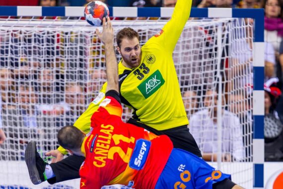 Ein Handballspieler in einem gelben Trikot (Torwart) blockiert einen Schuss, während ein Spieler in einem rot-blauen Trikot versucht, ein Tor zu erzielen; beide sind während eines Spiels in der Nähe des Tors in Aktion. Im Hintergrund sind Zuschauer zu sehen.