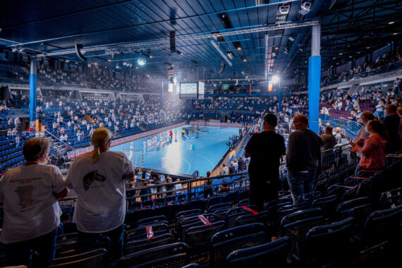Die Zuschauer in einer Halle verfolgen ein Handballspiel auf einem blauen Spielfeld, mit Teammitgliedern und Offiziellen auf dem Spielfeld und mit Zuschauern auf den Tribünen unter hellen Lichtern.