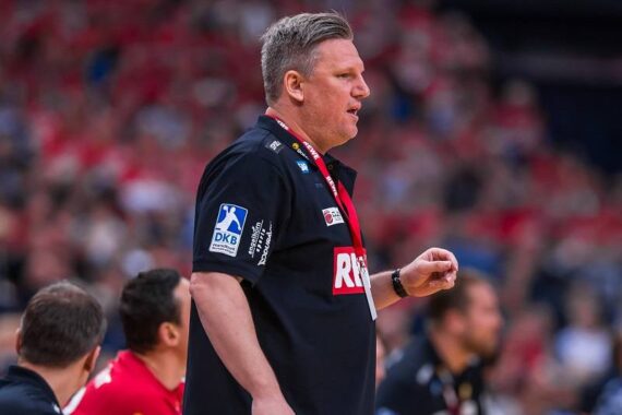 Ein Handballtrainer, der ein schwarzes Hemd mit Sponsorenlogos trägt, steht an der Seitenlinie und gibt während eines Spiels Anweisungen. Im Hintergrund ist ein verschwommenes Publikum zu sehen, das überwiegend in Rot gekleidet ist.