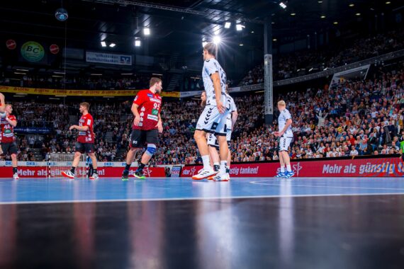 Handballspieler in rot-weißen Trikots spielen in einer großen Halle mit einer jubelnden Menge auf der Tribüne und hellen Lichtern über dem Kopf. Das Bild ist aus einem niedrigen Winkel in der Nähe des Spielfelds aufgenommen.