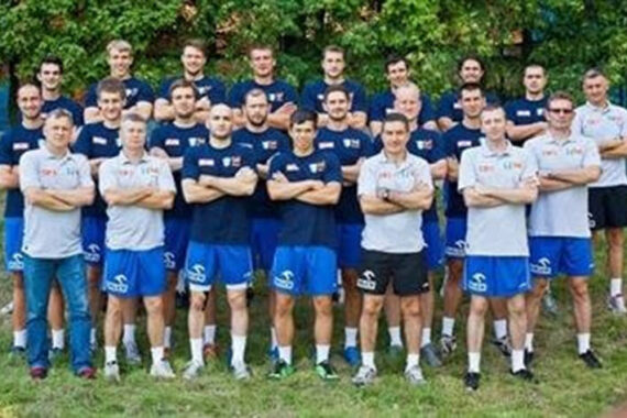 Eine Gruppe von männlichen Sportlern in passenden Sportuniformen und Trainern in weißen Hemden steht gemeinsam im Freien auf einer Wiese, die Arme verschränkt, und posiert für ein Mannschaftsfoto vor einem Baum.