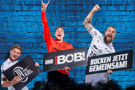 Drei aufgeregte Männer in Sporttrikots halten Schilder vor einer blauen Backsteinmauer hoch. Auf den Schildern steht THW Kiel, Und Radio Bob! und Rocken Jetzt Gemeinsam!.
