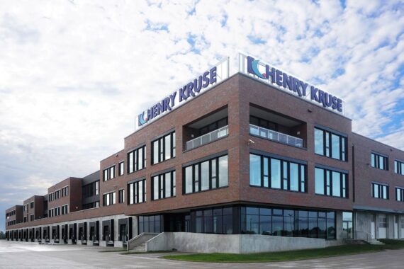 Ein großes, modernes Backstein-Bürogebäude mit Henry-Kruse-Schriftzug auf dem Dach, mehreren Fenstern und einer großen gepflasterten Fläche davor unter einem teilweise bewölkten Himmel.