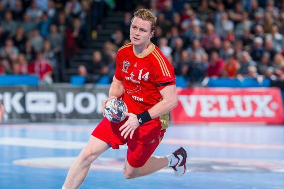 Ein männlicher Handballspieler in einem roten Trikot mit der Nummer 14 läuft auf einem Hallenplatz und hält einen Handball, während im Hintergrund Zuschauer und Werbung zu sehen sind.
