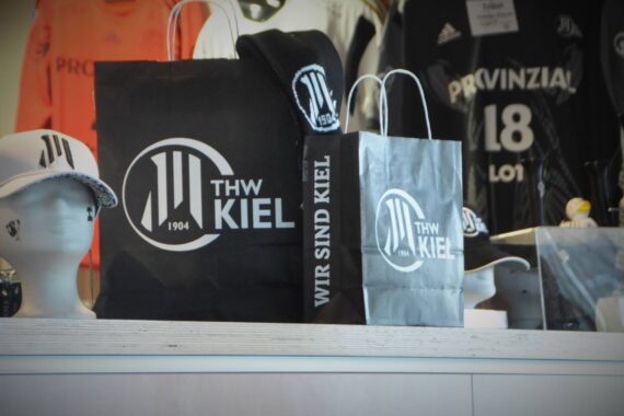 Zwei schwarze Einkaufstaschen mit dem Logo des THW Kiel liegen auf einer Theke, daneben weiße Mützen auf Schaufensterköpfen und schwarze Sporttrikots im Hintergrund. Die Szene scheint sich in einem Warenhaus zu befinden.