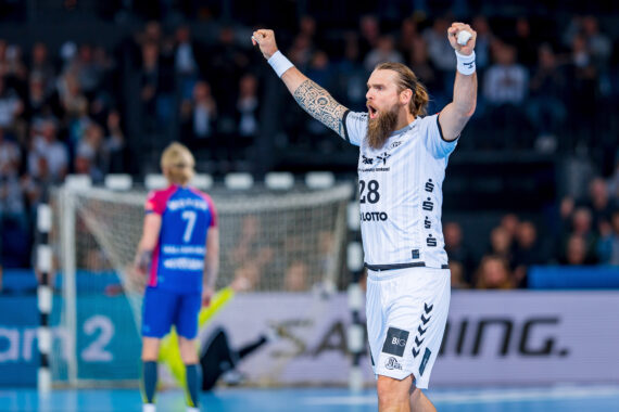 Ein bärtiger Handballspieler in einem weißen Trikot hebt jubelnd die Arme auf dem Spielfeld, während ein anderer Spieler in einem blau-rosa Trikot in der Nähe des Tores im Hintergrund steht.