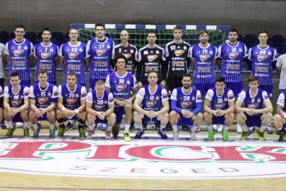 Eine Männerhandballmannschaft posiert in einer Halle auf einem Spielfeld. Sie tragen blau-weiße Trikots mit dem Aufdruck "PICK" und Sponsoren. Einige stehen, andere knien vor einem Tor, auf dessen Boden ein großes "PICK SZEGED"-Logo prangt.