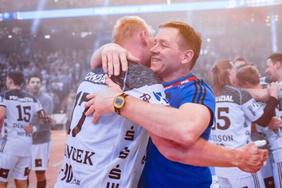 Ein Trainer in einem blauen Hemd umarmt freudig einen Spieler in einem weiß-schwarzen Trikot auf einem überfüllten Hallenplatz, während seine Mannschaftskameraden im Hintergrund jubeln.