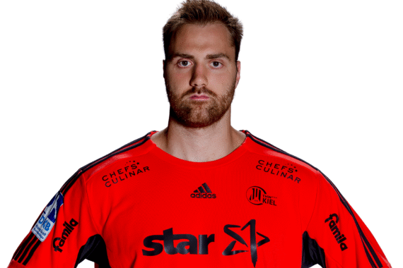 Ein Mann mit kurzen braunen Haaren und einem Bart trägt ein leuchtend rotes Sporttrikot mit Sponsorenlogos. Er steht vor einem weißen Hintergrund und blickt mit ernster Miene direkt in die Kamera.