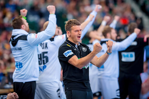 Ein Mann in einem schwarzen Hemd reckt die Fäuste in die Höhe, während im Hintergrund mehrere Personen in weißer und schwarzer Sportkleidung bei einem Sportereignis begeistert jubeln.