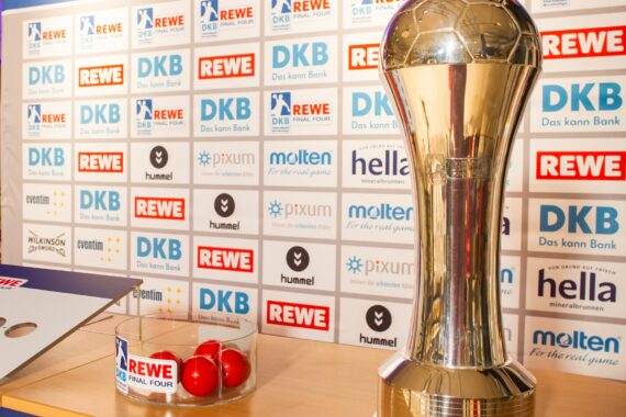 Ein großer, glänzender Pokal steht auf einem Tisch neben einer Schale mit roten Bällen, vor einem Hintergrund, der mit verschiedenen Sponsorenlogos wie REWE, DKB und hummel geschmückt ist.