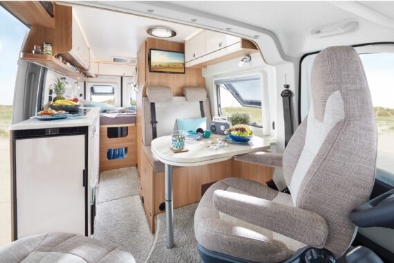 Der Innenraum eines Wohnmobils besteht aus einem Esstisch mit Speisen und Getränken, gepolsterten Sitzen, einem Bett im Fond, einer kleinen Küchenzeile, hellen Holzschränken und einem Fenster mit Blick auf die Sanddünen draußen.