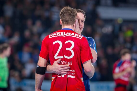 Ein Mann in einem blauen Hemd tröstet einen Handballspieler, der ein rotes Trikot mit der Nummer 23 trägt, indem er während eines Spiels in einer Halle seine Hand auf das Gesicht des Spielers legt. Andere Spieler sind im unscharfen Hintergrund zu sehen.