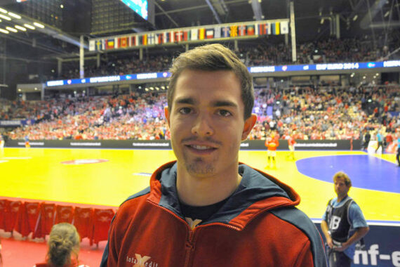 Ein junger Mann in einer roten Jacke steht in einer überfüllten Sportarena mit einem Handballfeld und vielen Zuschauern im Hintergrund. Die Halle ist hell erleuchtet, und über dem Kopf hängen Nationalflaggen.