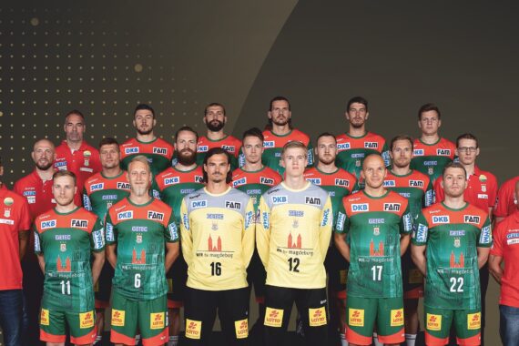 Eine Herrenhandballmannschaft stellt sich für ein Gruppenfoto auf. Die Spieler tragen grüne und gelbe Trikots mit Logos, und die Trainer in roten Hemden stehen an beiden Enden und in der hinteren Reihe. Der Hintergrund ist dunkel mit goldenen Punkten und geometrischen Formen.