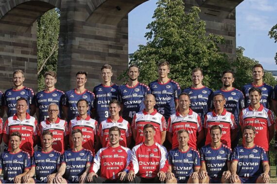 Ein Gruppenfoto eines Herrensportteams in blauen und roten Uniformen, das im Freien in Reihen posiert, mit Bäumen und Steinbögen im Hintergrund.