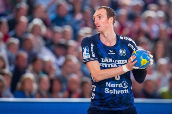Ein männlicher Handballspieler in einem blauen Trikot hält einen bunten Handball und blickt während eines Spiels nach links, während im Hintergrund eine unscharfe Menschenmenge zu sehen ist.