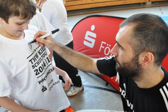 Ein Mann mit Bart signiert ein weißes T-Shirt, das von einem lächelnden Kind bei einer Indoor-Veranstaltung getragen wird. Auf dem T-Shirt sind Text und Logos aufgedruckt. Im Hintergrund ist ein rotes Werbebanner zu sehen.