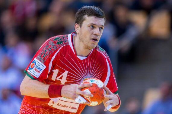Ein männlicher Handballspieler in einem roten Trikot mit der Nummer 14 hält einen Ball und bereitet sich auf den Wurf während eines Spiels vor. Der Hintergrund ist unscharf und konzentriert sich auf seinen entschlossenen Ausdruck und seine Aktion.