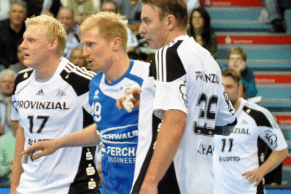 Drei männliche Handballspieler in Aktion während eines Spiels, zwei in weißen Trikots und einer in einem blauen Trikot, mit einer unscharfen Zuschauermenge und Stadionbestuhlung im Hintergrund.