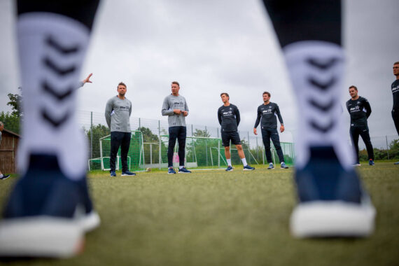 Eine Gruppe von Männern in Sportkleidung steht in einem Halbkreis auf einem Sportplatz, gesehen durch die Beine einer Person im Vordergrund, die weiße Socken und blaue Turnschuhe trägt. Der Himmel ist bedeckt.