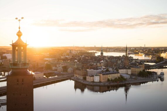 Luftaufnahme von Stockholm bei Sonnenaufgang, mit von Wasser umgebenen Gebäuden, Brücken und dem markanten Turm des Stockholmer Rathauses im Vordergrund.