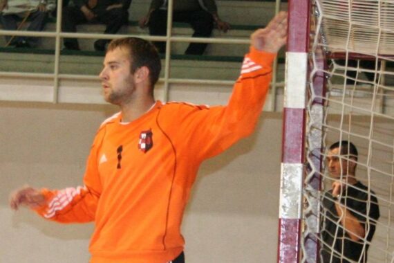 Ein Handballtorwart in einem orangefarbenen Trikot steht mit erhobenem Arm in der Nähe des Torpfostens und wirkt während eines Spiels konzentriert. Im Hintergrund sind Zuschauer zu sehen.