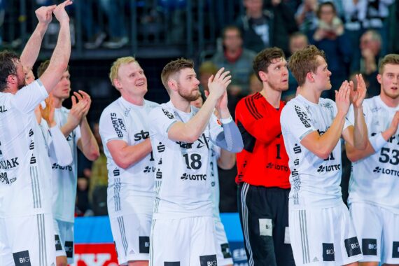 Eine Gruppe von männlichen Handballspielern in weißen Trikots und einer in einem roten Trikot stehen zusammen, klatschen und schauen nach vorne, im Hintergrund eine Menschenmenge.