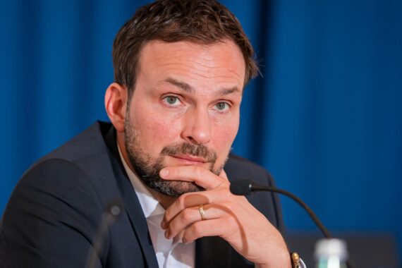 Ein Mann mit kurzen braunen Haaren und einem Bart, der einen dunklen Anzug und ein weißes Hemd trägt, sitzt an einem Tisch mit Mikrofonen, stützt sein Kinn auf die Hand und blickt nachdenklich nach vorn. Im Hintergrund ist ein blauer Vorhang zu sehen.