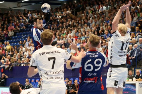 Ein Handballspieler in einem blau-roten Trikot springt, um den Ball zu schießen, während drei Verteidiger in weißen Trikots ihn abwehren. Das Spiel findet in einer überfüllten Halle statt, in der die Zuschauer aufmerksam zusehen.