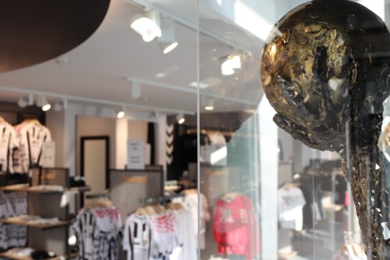 Nahaufnahme einer bronzenen Fußballtrophäe in einer Glasvitrine, im Hintergrund ein Sportartikelgeschäft mit schwarzen und weißen Trikots und Shirts auf Regalen.
