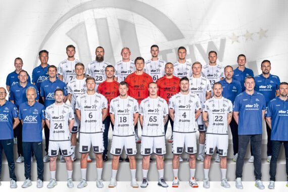 Eine Herren-Handballmannschaft stellt sich für ein offizielles Gruppenfoto auf. Die Spieler in weißen Trikots und die Trainer in blauen Hemden stehen auf einer Treppe vor einem hellen Hintergrund mit einem schwachen Logo.