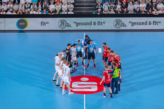 Zwei Handballmannschaften, eine in Weiß und eine in Rot, stehen vor einem Spiel in einem Kreis mit den Schiedsrichtern in der Mitte des Spielfelds, umgeben von Zuschauern in einer hell erleuchteten Halle.